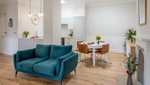 A modern living room with a teal sofa, a dining area with a white table and brown chairs, and a kitchen in the background. - The Swaledale -  a cottage that sleeps 2 guests  in 1 bedroom (Richmond)