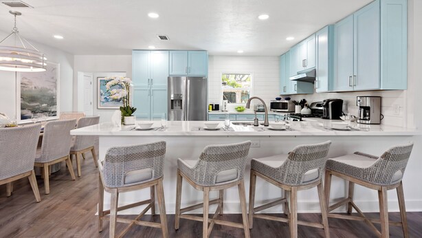 A modern kitchen with light blue cabinets, a large island with bar stools, stainless steel appliances, and a dining area with wicker chairs. - Tropical Oasis with Private Pool & Spa, minutes from Siesta Key Beach! (Sarasota)