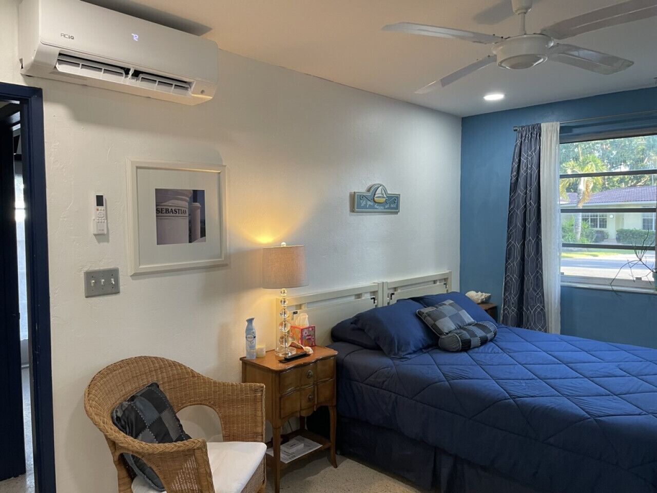 Ein Schlafzimmer mit einem Bett, einem Nachttisch, einem Sessel, einem Fenster mit Vorhängen und einem Deckenventilator.