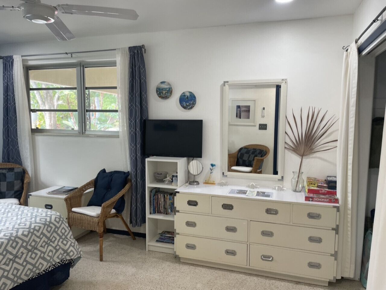 Un dormitorio con una cama, una silla, un tocador, un televisor y una ventana con cortinas.