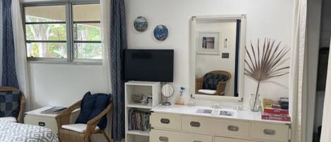 Un dormitorio con una cama, una silla, un tocador, un televisor y una ventana con cortinas.