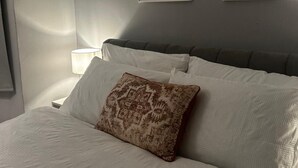 Ein ordentlich bezogenes Bett mit weißen Leinentüchern und einem gemusterten Kopfkissen, eine Nachttischlampe und zwei gerahmte Bilder an der Wand.