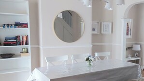 Ein Essbereich mit einem Tisch, der mit einer weißen Tischdecke bedeckt ist, umgeben von weißen Stühlen, einem runden Spiegel an der Wand und einem Bücherregal mit Büchern und Dekoration.