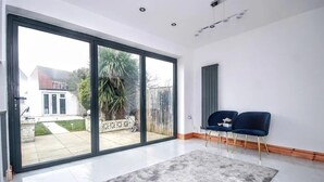 Ein geräumiges Zimmer mit einem großen Schiebeglasfenster, das auf eine Terrasse führt, zwei blaue Stühle und ein grauer Teppich.