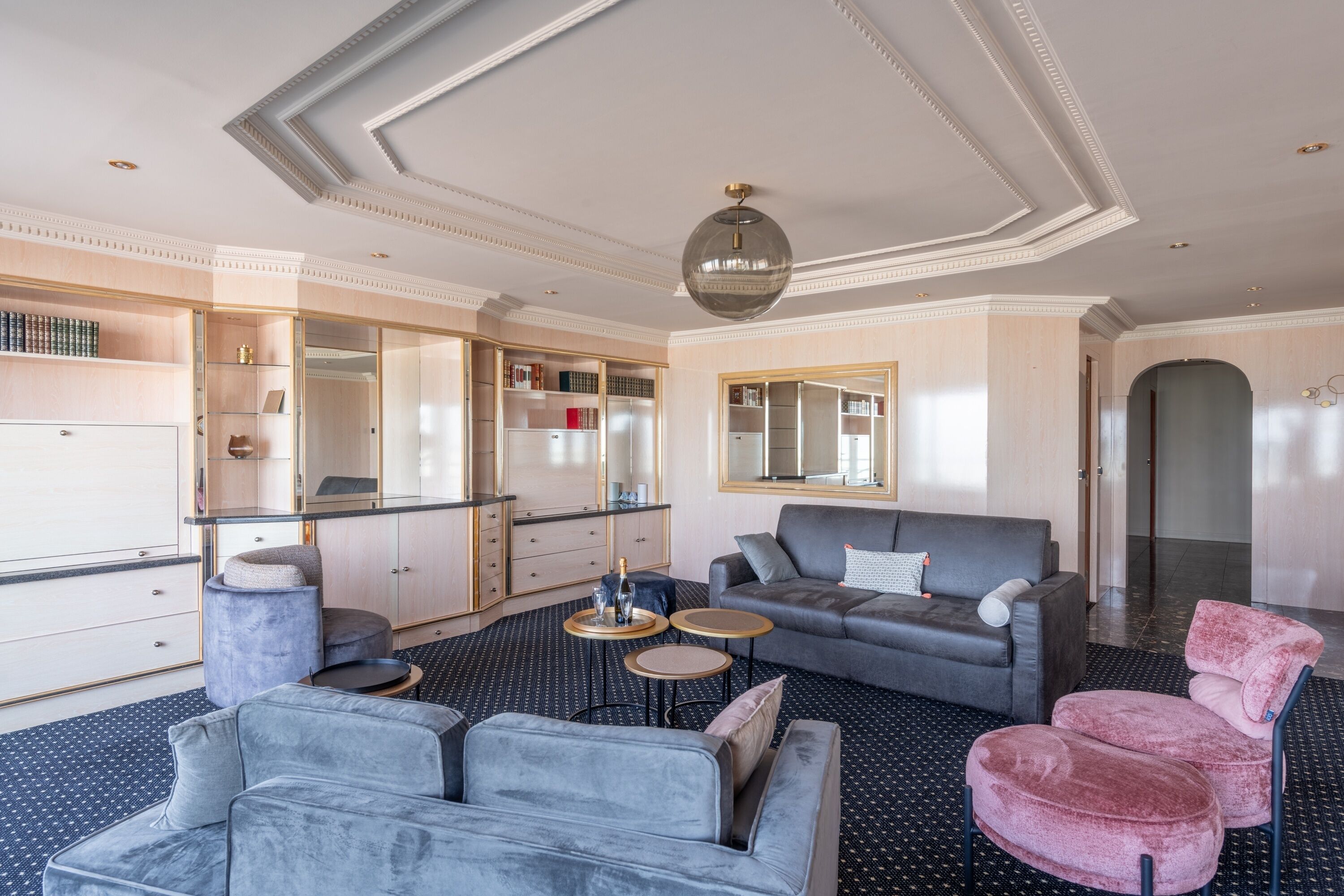 Una camera d'albergo moderna con un divano grigio, una poltrona rosa, un tavolino rotondo e un grande specchio.