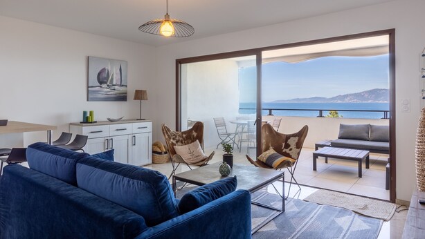 Un salon moderne avec un canapé bleu, une table basse et un coin repas. Une grande fenêtre offre une vue sur la mer et les montagnes.