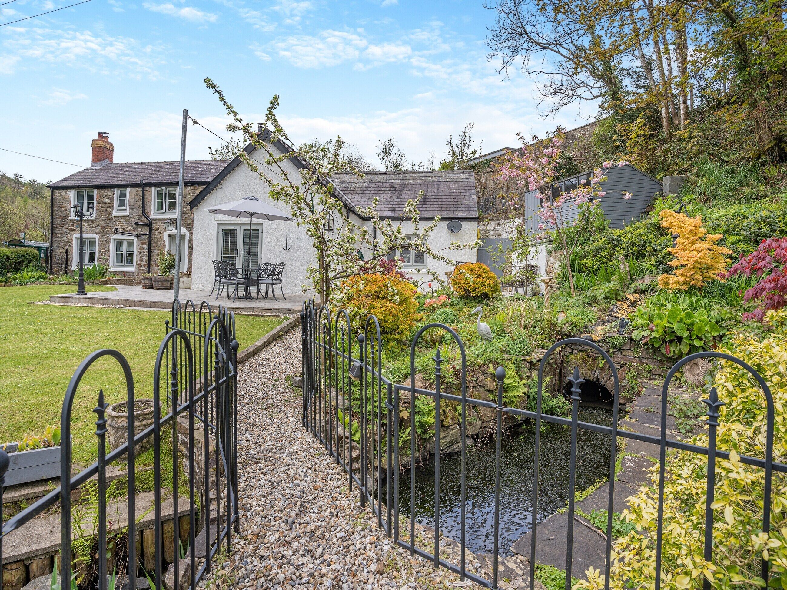 A stone house with a garden, a small stream, and a metal fence.