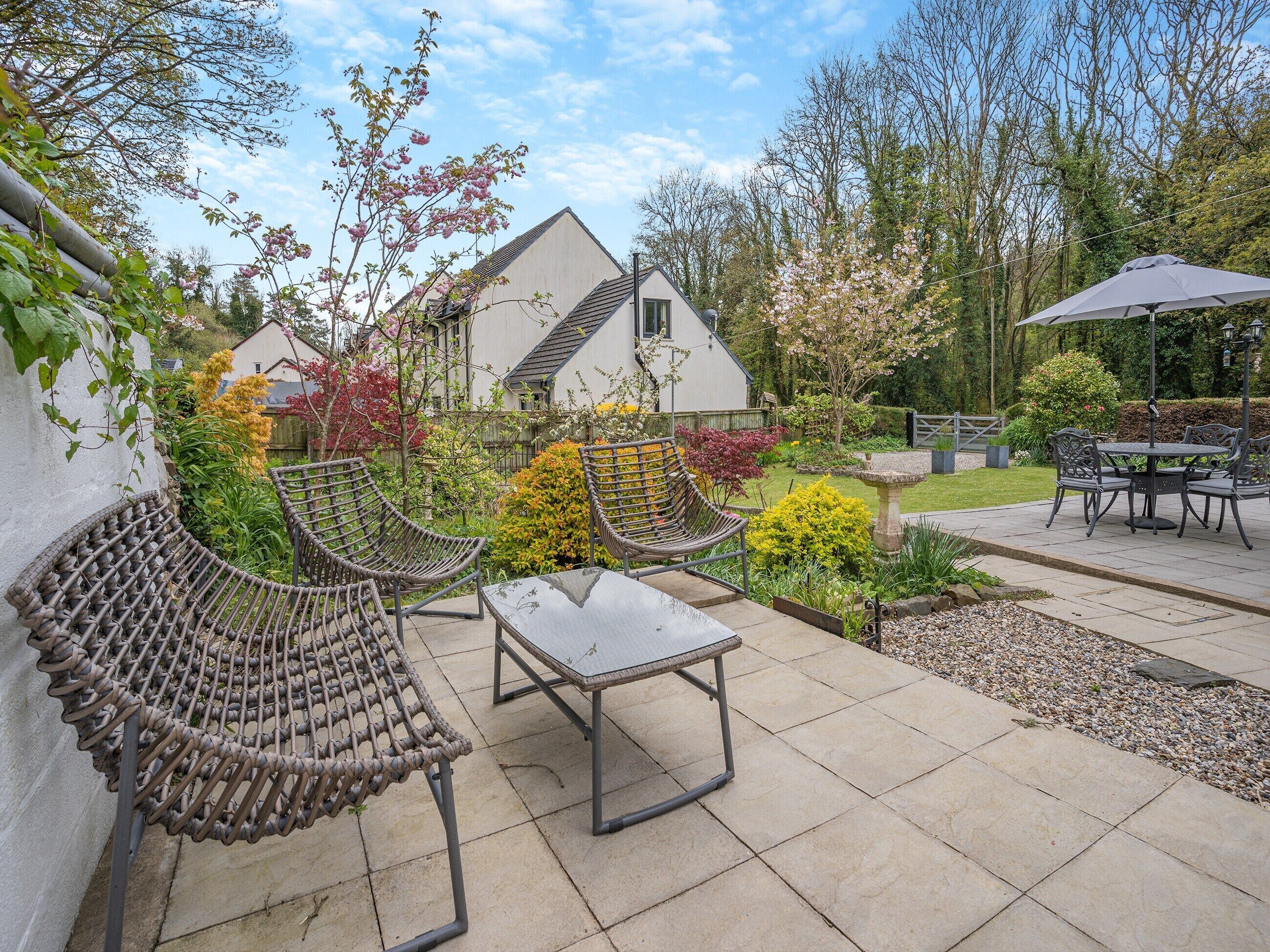 A patio with wicker chairs and a table, surrounded by a garden with various plants and a house in the background.