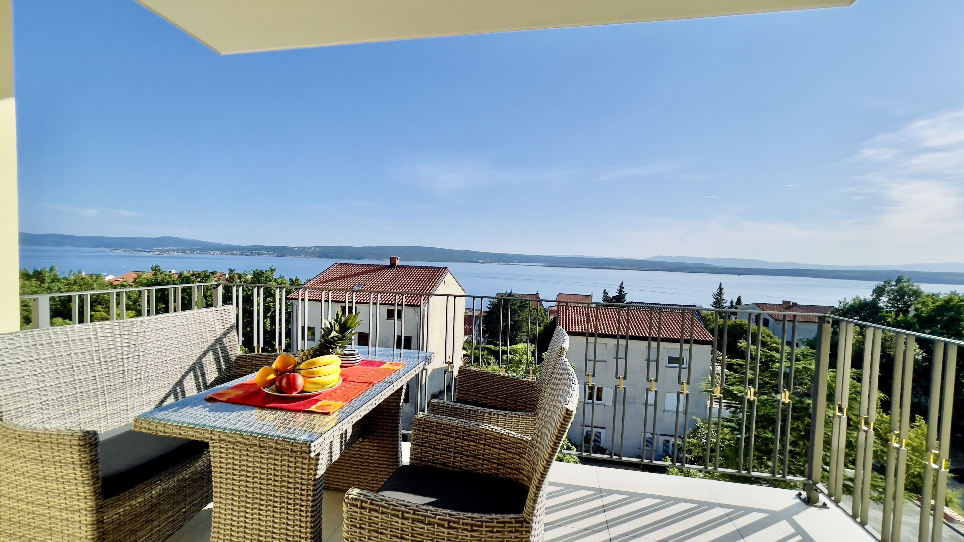 A balcony with wicker furniture, a table with fruit, and a view of a lake and buildings.