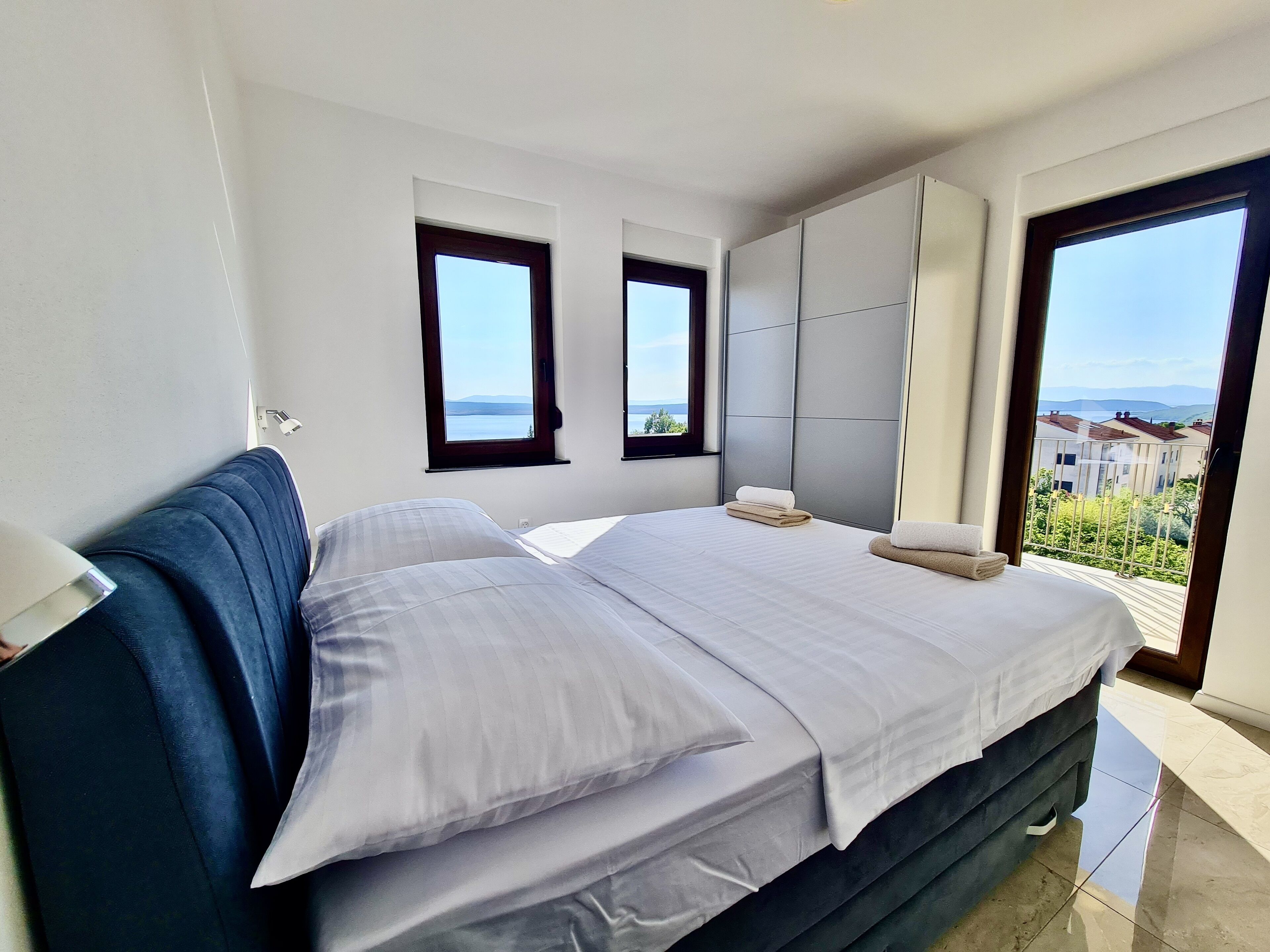 A bedroom with a large bed, two windows, a wardrobe, and a view of the sea.