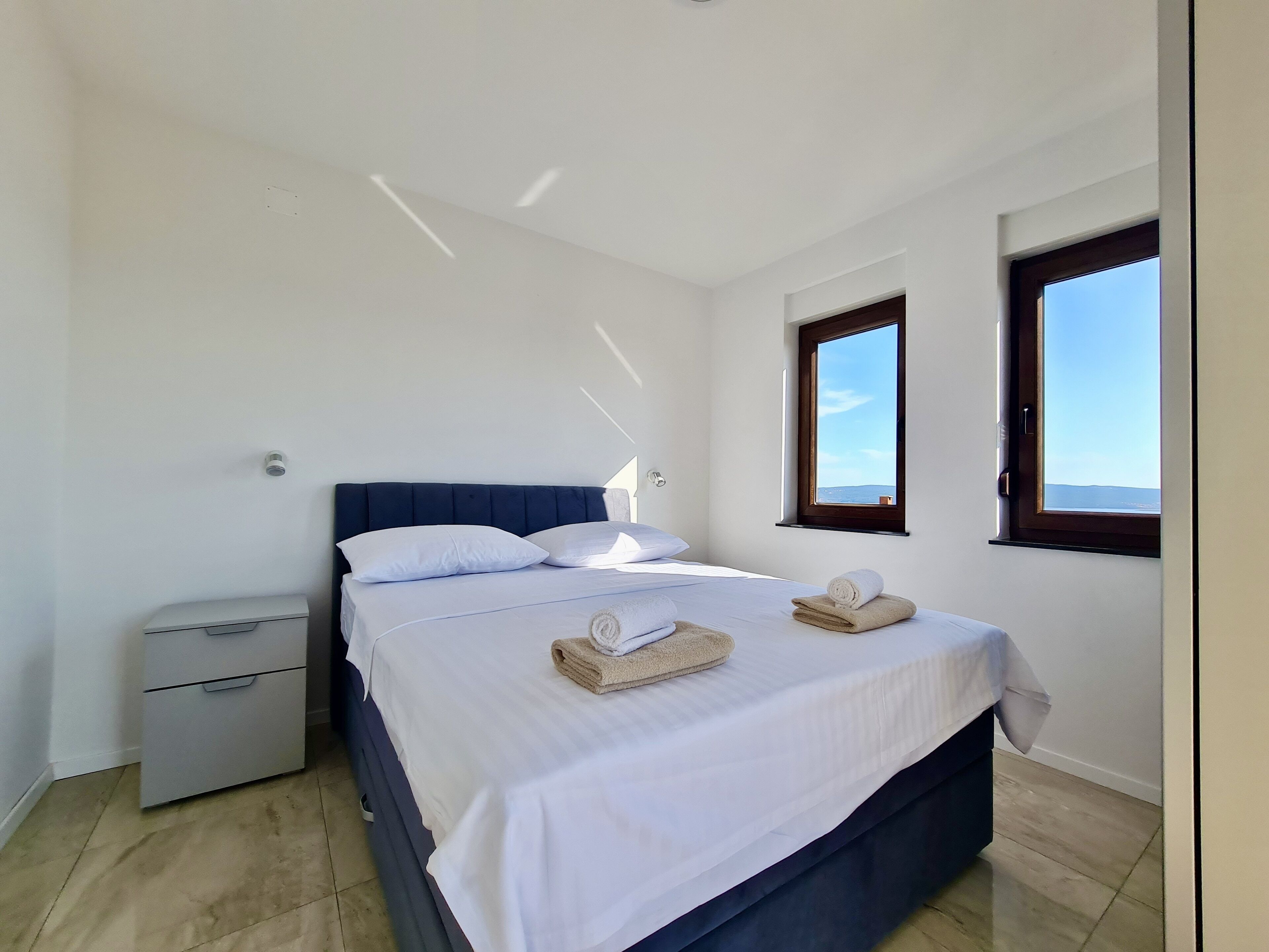 A bedroom with a bed, two windows, a nightstand, and a drawer unit.