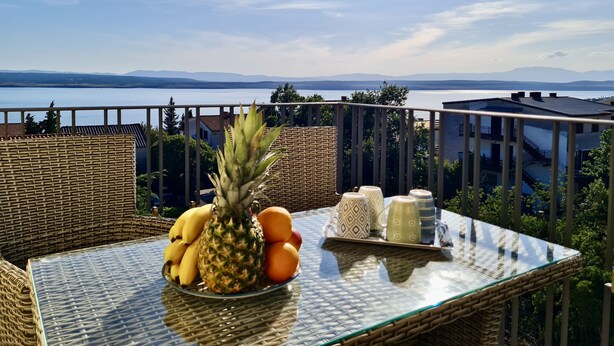 Ein Korbtisch mit Glasplatte, eine Fruchtschale und ein Tablett mit Tassen und Gläsern auf einem Balkon mit Blick auf einen See und Berge.