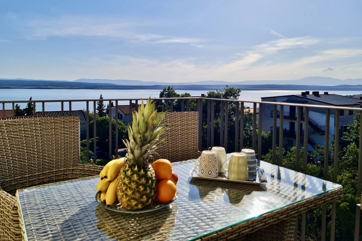 A wicker table with a glass top, a plate of fruit, and a tray with cups and jars on a balcony overlooking a lake and mountains.