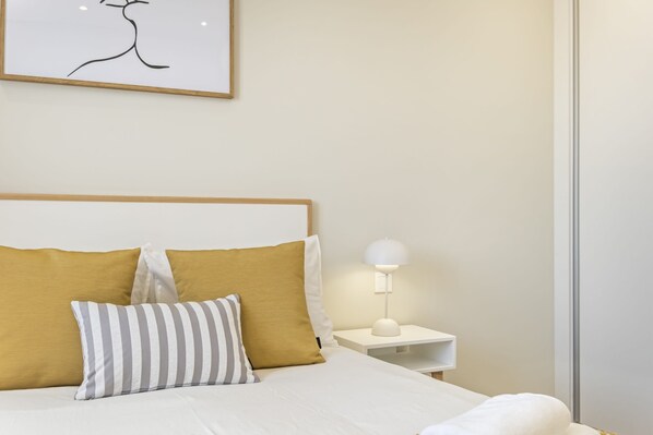 A neatly made bed with a white comforter and two pillows, one striped and one solid yellow. A white bedside lamp is on the nightstand beside the bed.