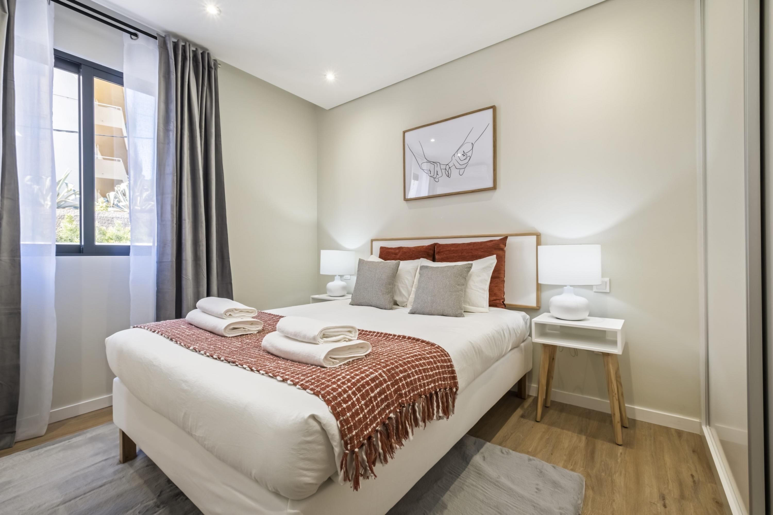 A modern hotel room with a large bed, two bedside tables, a window with curtains, and a framed artwork on the wall.