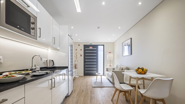 A modern kitchen with white cabinets, a black countertop, and a built-in microwave. There is a dining area with a white table and chairs, and a small rug on the floor.
