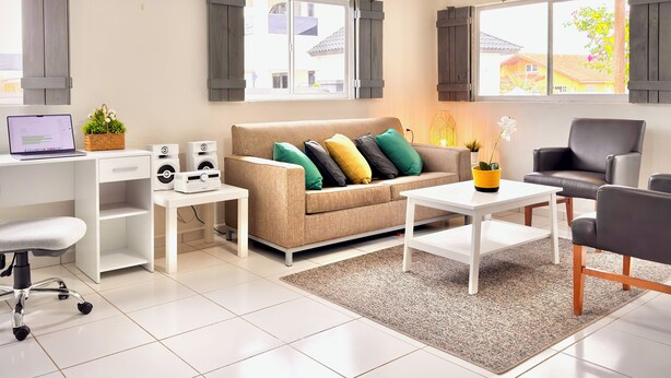 En moderne stue med en beige sofa, et hvidt sofabord, en sort læderstol, et hvidt skrivebord med en bærbar computer, højttalere og en lille plante.