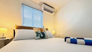 Et hotelværelse med en seng, to sengeborde med lamper, et vindue med persienner, en aircondition og en pyntende pude.
