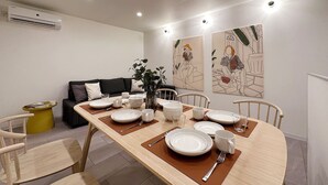 A dining area with a wooden table set for four, surrounded by chairs. There is a sofa and a small yellow side table in the background. - Nikke 202 (Hiroshima)