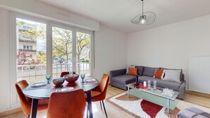 A modern living room with a dining area, large windows, a grey sofa, and a glass coffee table. - Brasseur (Colmar)