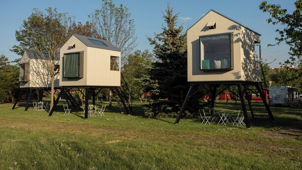 Two elevated treehouse-style cabins with open sides, set on stilts in a grassy area with trees and a clear sky. - Camp Dzban (Prague)