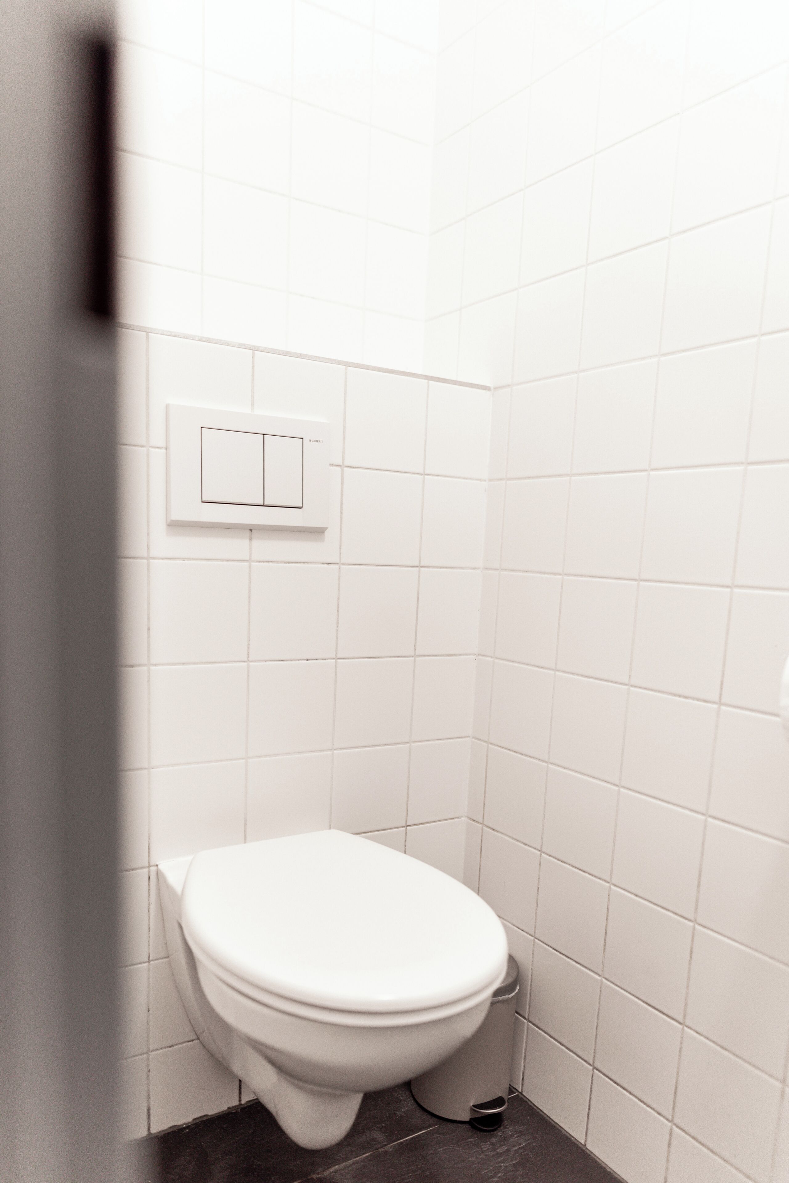 Et lille badeværelse med hvide fliser, et hvidt toilet, en lyskontakt på væggen og en skraldespand på gulvet.