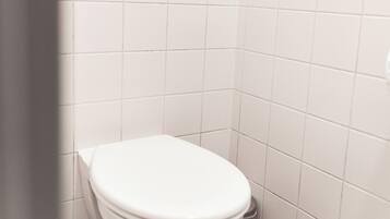 Et lille badeværelse med hvide fliser, et hvidt toilet, en lyskontakt på væggen og en skraldespand på gulvet.