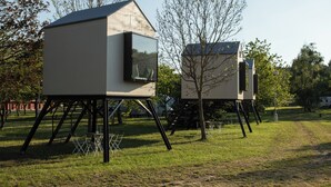 Two modern, elevated structures with large windows, set in a grassy area with trees and a clear sky. - Camp Dzban (Prague)