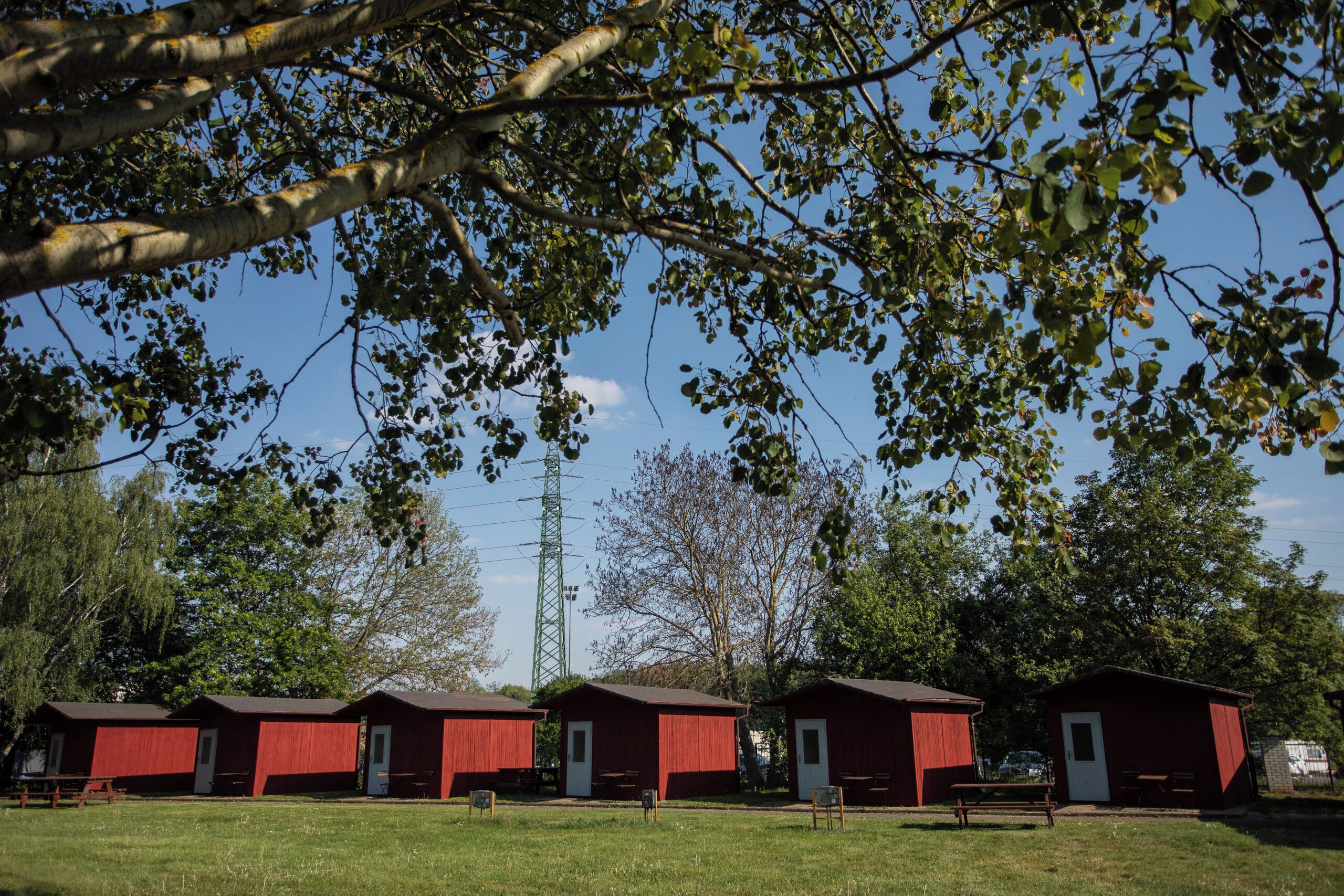 A row of red wooden cabins with white doors, set in a grassy field with trees and a clear sky in the background.