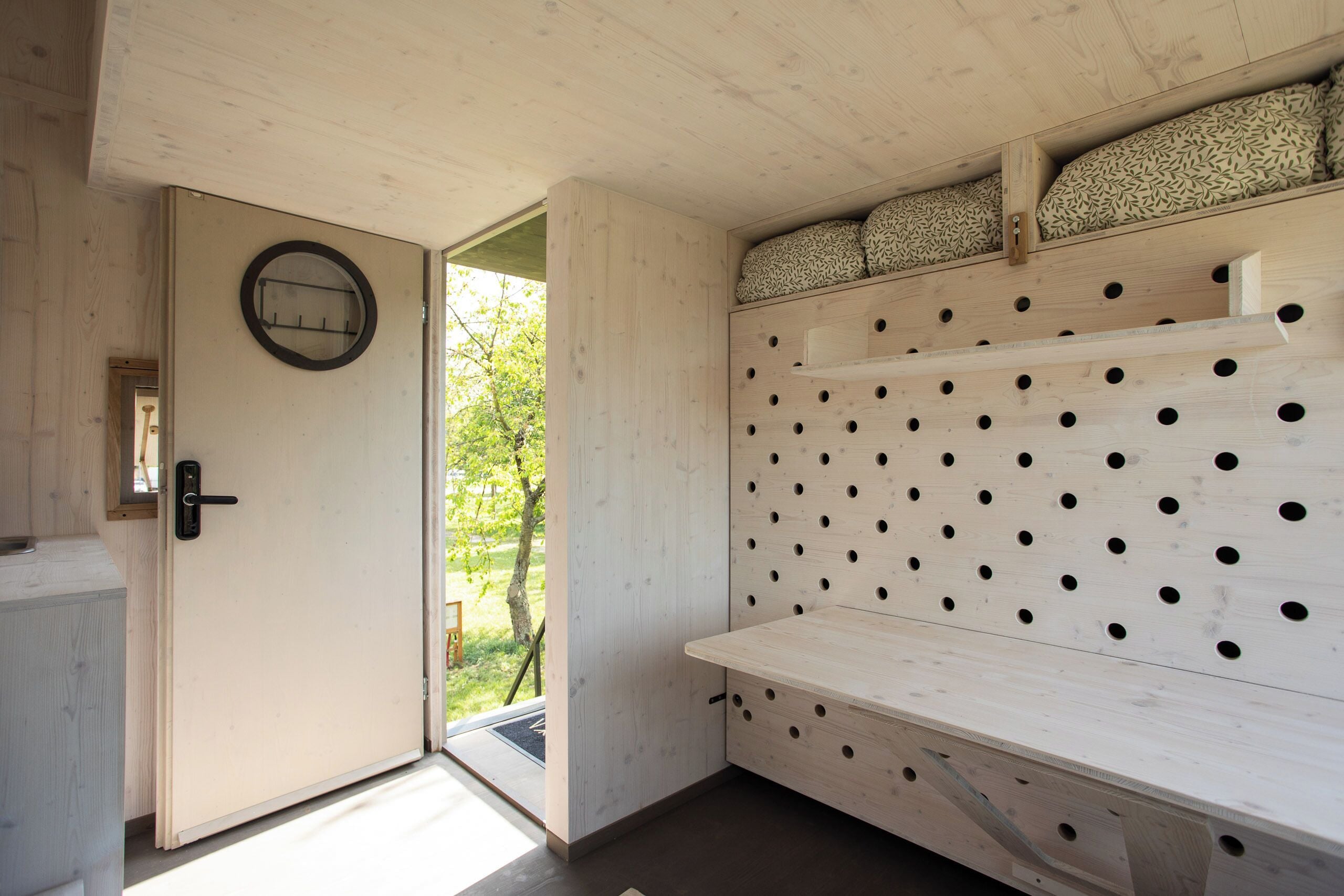 A wooden room with a door, a wall-mounted shelf with patterned cushions, and a bench with circular cutouts.