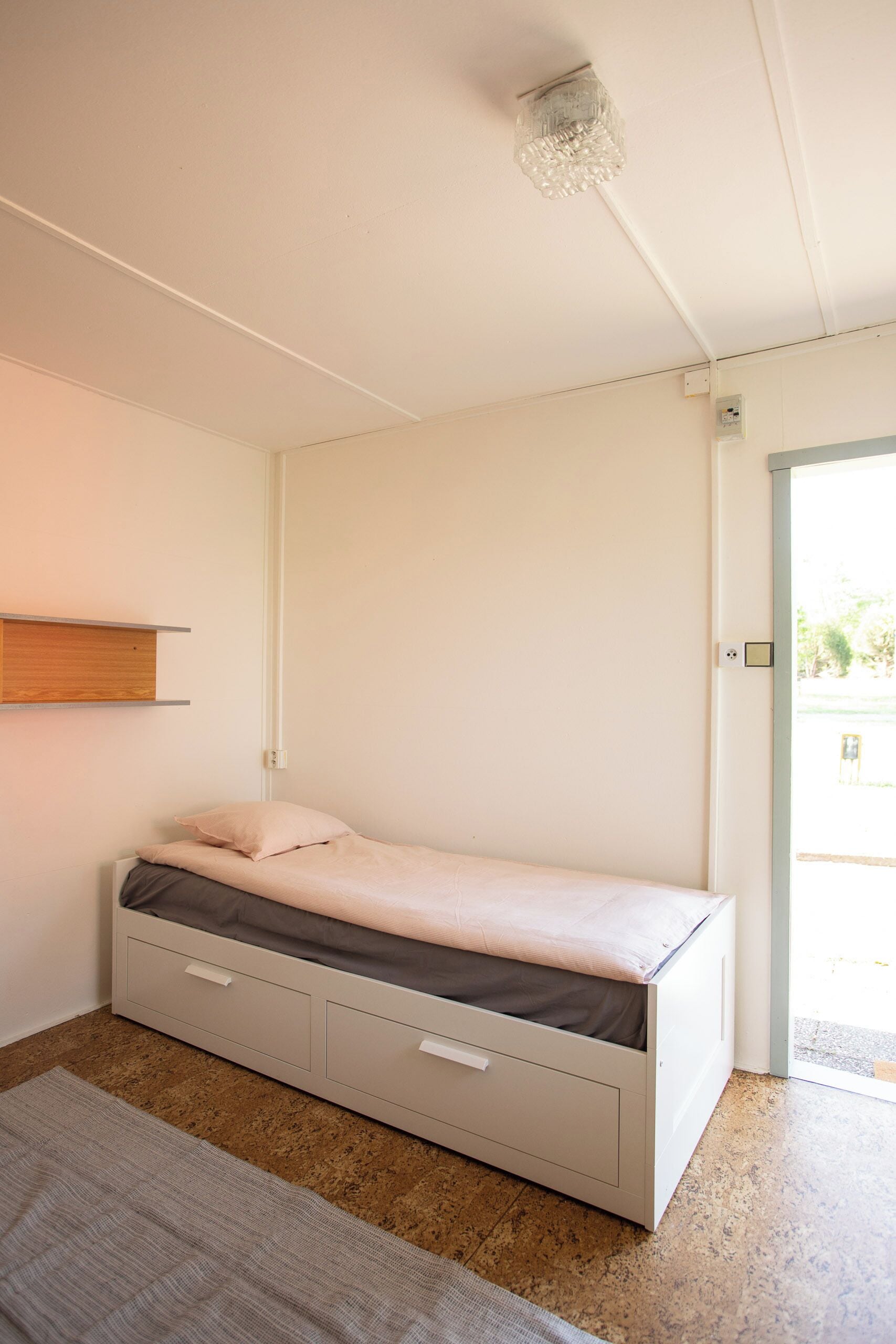 A minimalist bedroom with a bed, a small shelf, and a door leading to a balcony.