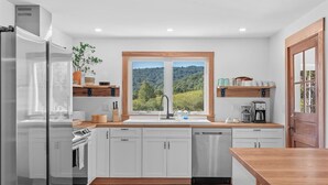 A modern kitchen with white cabinetry, a stainless steel refrigerator, a dishwasher, and a window with a view of greenery. - Secluded Cottage on 85 Acres – Peaceful Country Retreat Surrounded by Nature (Walton)