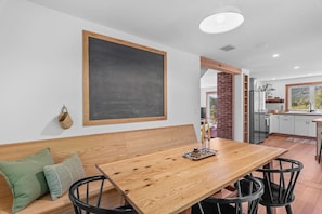 Un comedor con una mesa de madera, sillas negras, un banco con cojines, una pizarra en la pared y una cocina visible a través de un vano abierto.