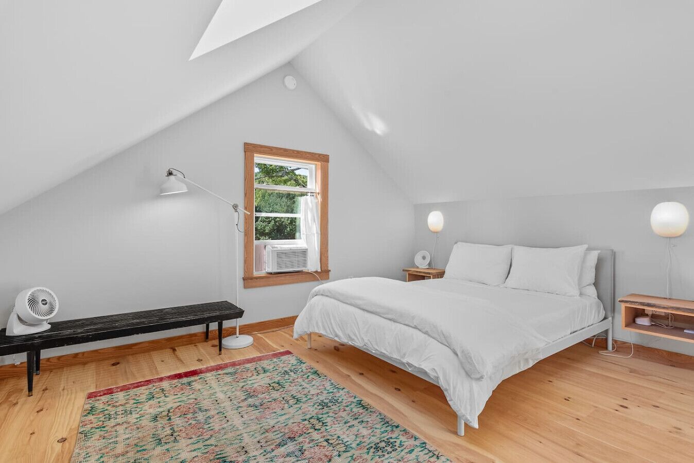 A bedroom with a sloped ceiling, a bed, a bench, a desk, and a window with an air conditioner.