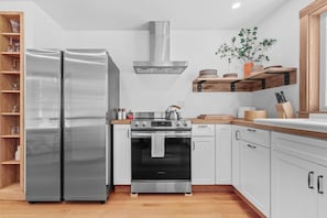 Una cocina moderna con refrigerador de acero inoxidable, estufa a gas, horno, gabinetes blancos, estantes de madera y una planta en maceta.
