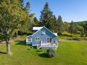 Une maison bleue avec un porche et une terrasse, entourée de pelouse verdoyante et d’arbres.