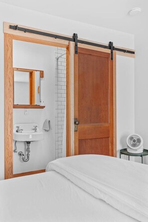 A bedroom with a bed, a wooden door, a bathroom with a sink, and a fan.