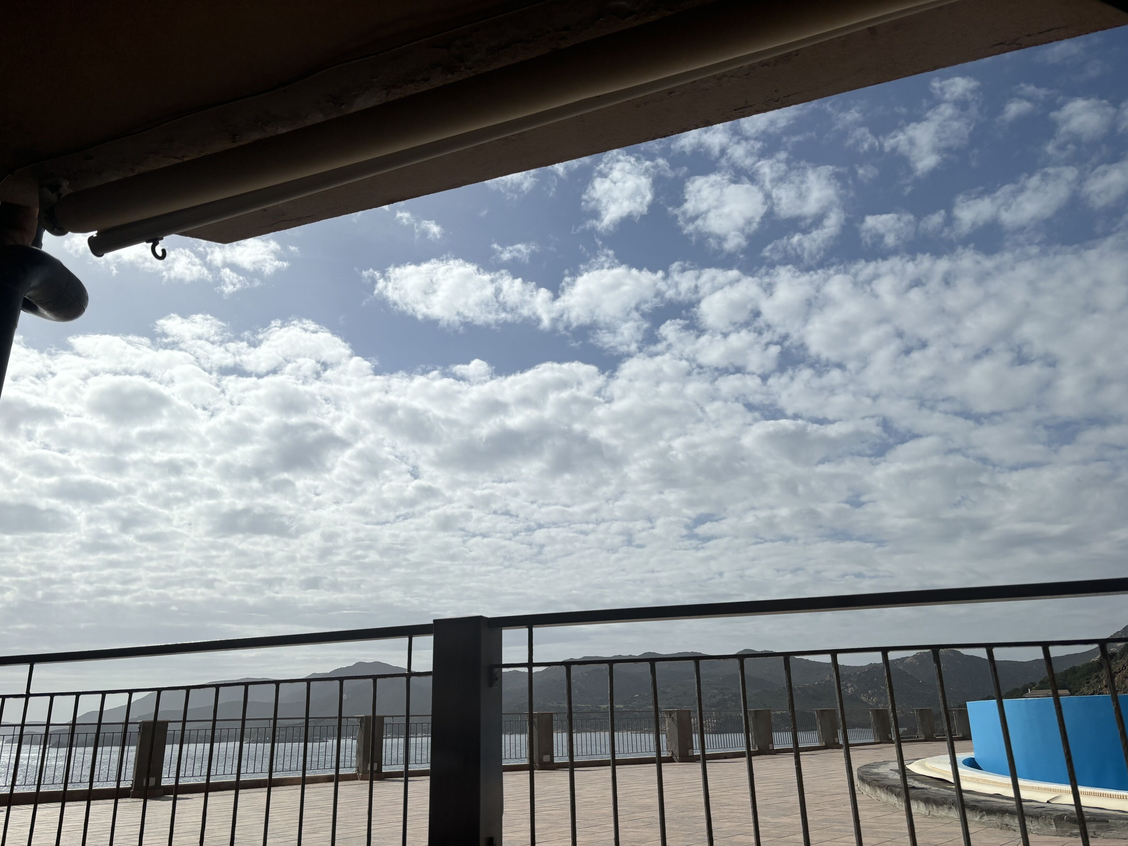 A view from a balcony with a metal railing, overlooking a body of water and distant mountains under a partly cloudy sky.