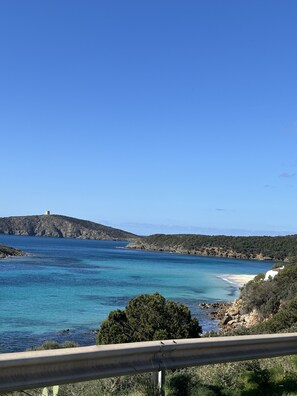 Paisaje costero con un faro en una colina, mar azul transparente y terreno rocoso.