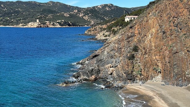 Un paysage côtier caractérisé par une eau d’un bleu limpide, une plage de sable, des falaises rocheuses et une structure en forme de tour située sur une colline.