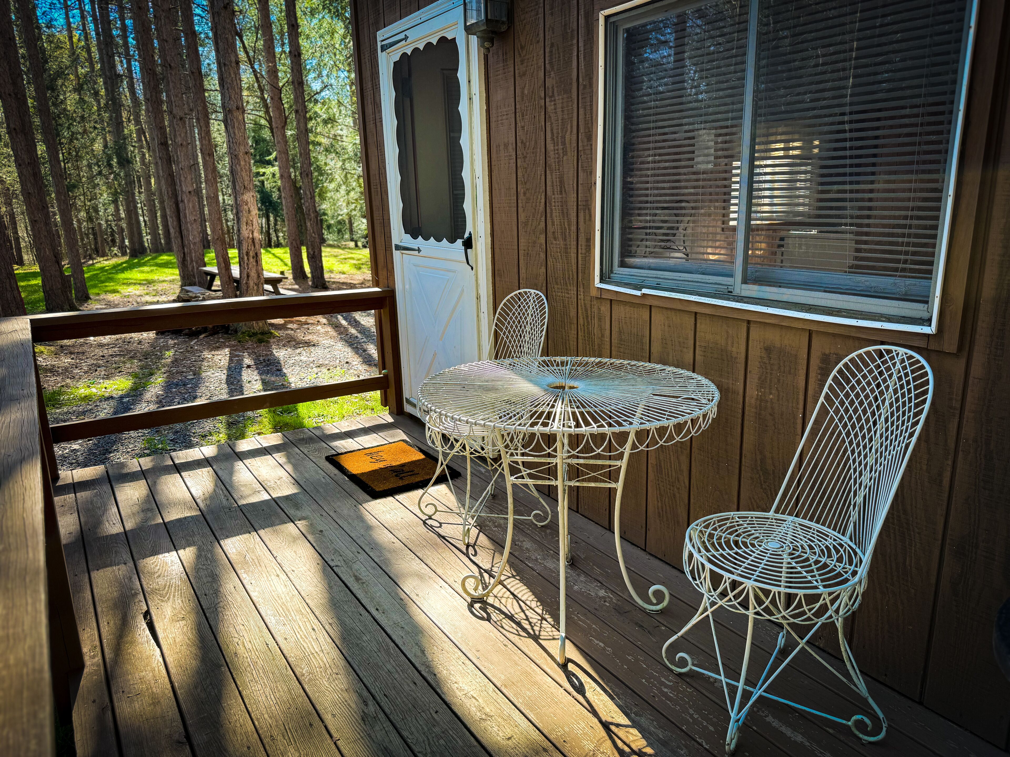 A wooden deck with a white metal table and two chairs, a door, and a window with blinds.