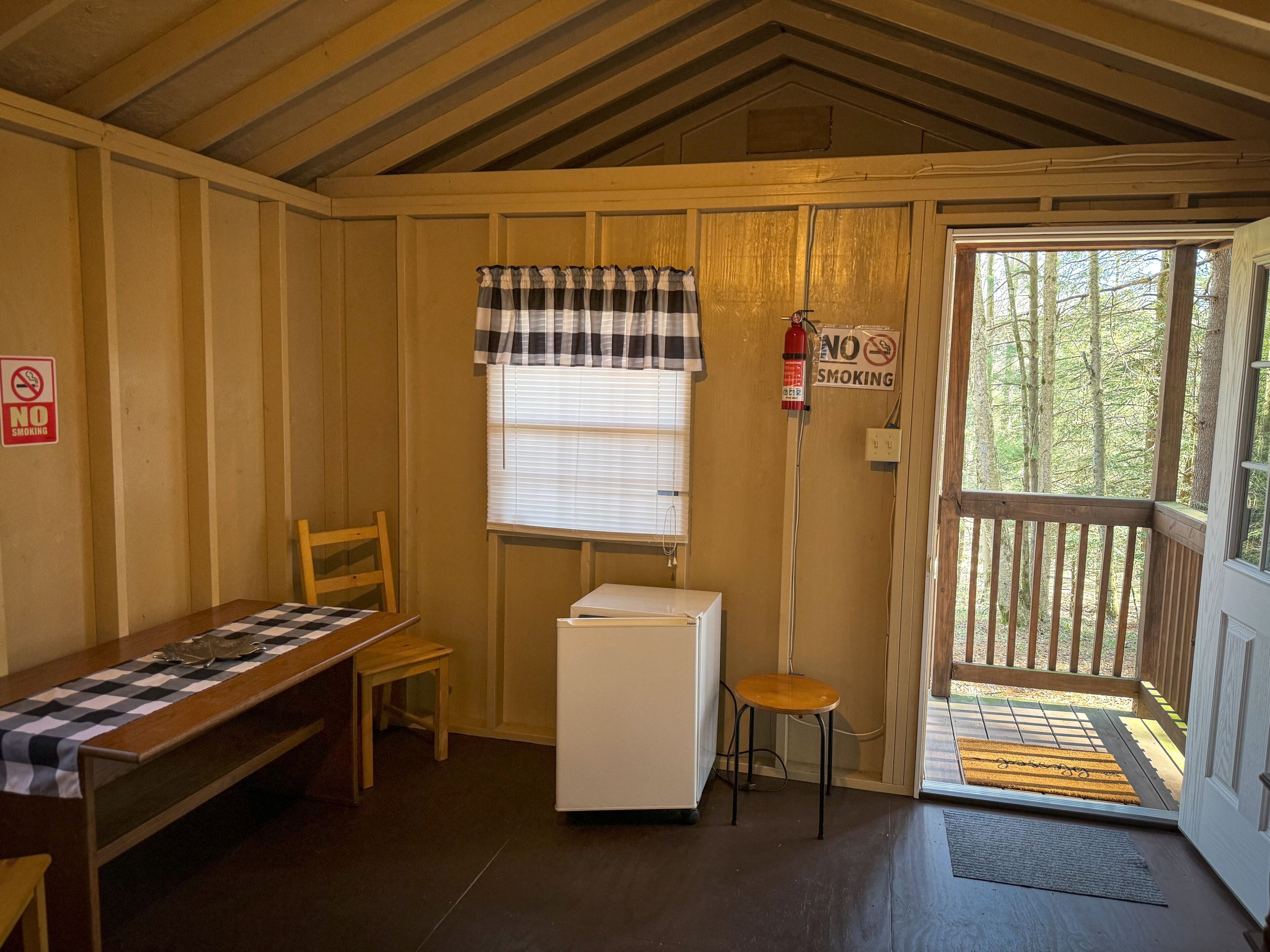 Una habitación pequeña y acogedora con un banco de madera, un refrigerador blanco y una ventana con persianas. Hay un cartel de 'No Fumar' en la pared y una puerta que da a una terraza al aire libre.