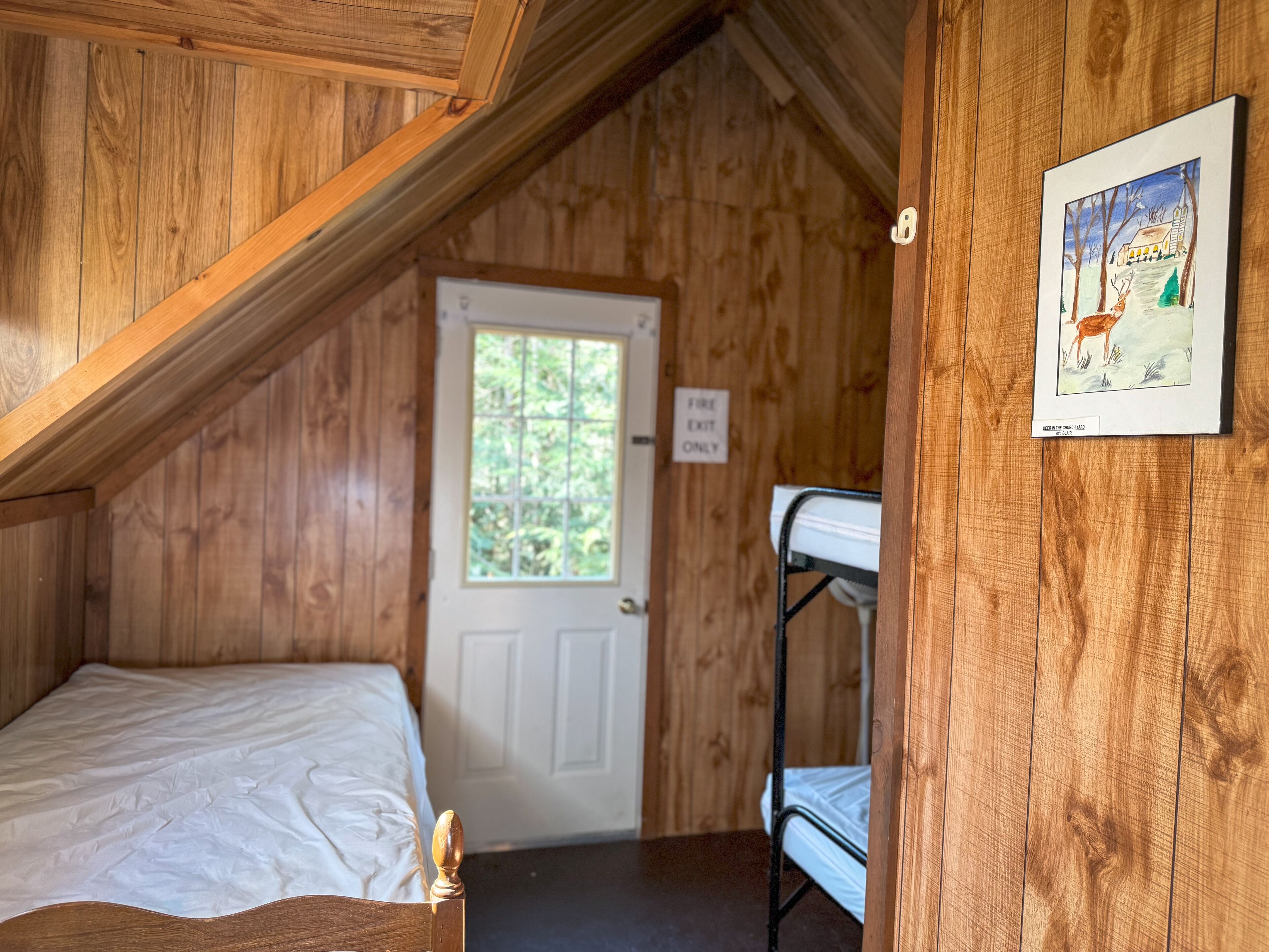 A wooden room with a bunk bed, a window, and a door marked "FIRE EXIT ONLY".