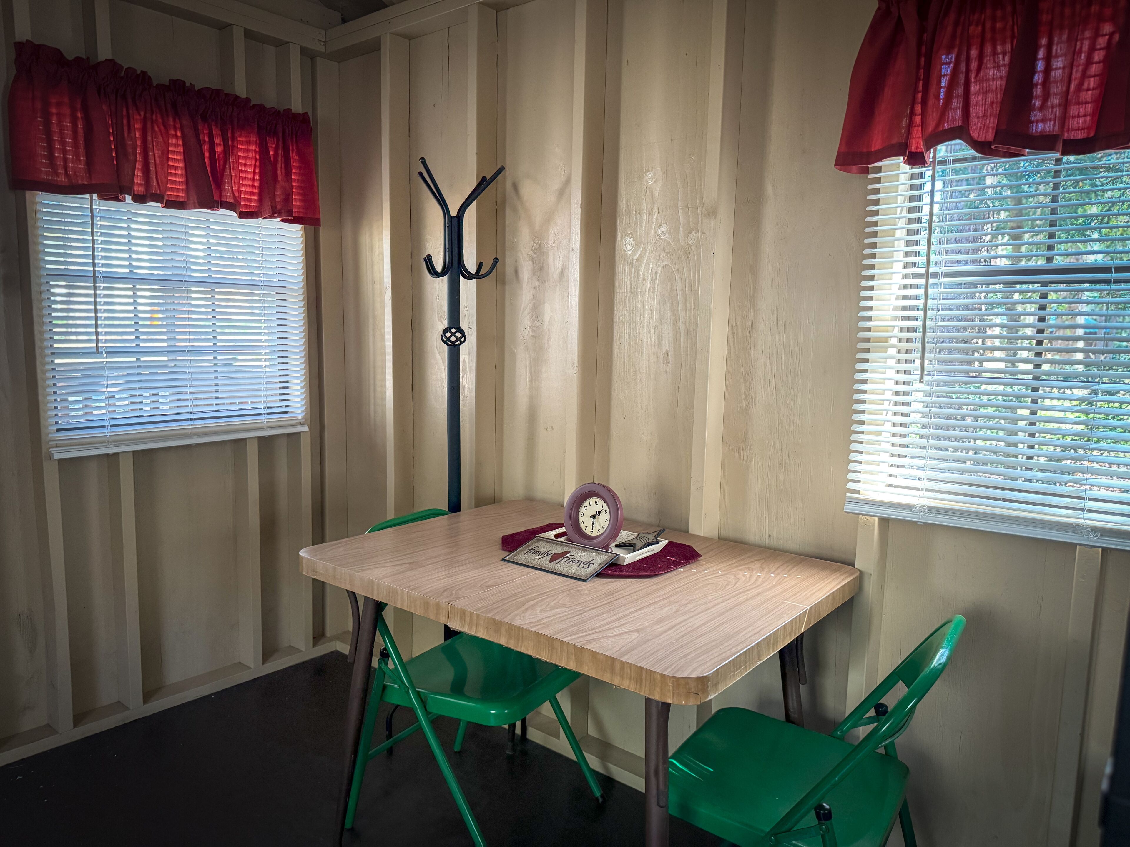 A room with a wooden table, green chairs, a coat rack, and two windows with blinds.