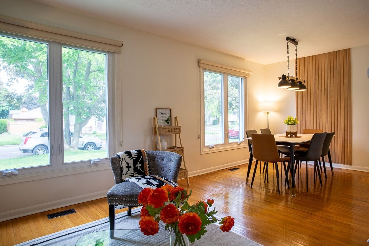 Uma sala de jantar com mesa e cadeiras, janelas amplas, uma poltrona com manta estampada e um vaso com flores.