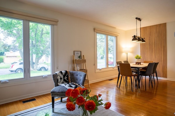 A dining area with a table and chairs, large windows, a chair with a patterned throw, and a vase with flowers.