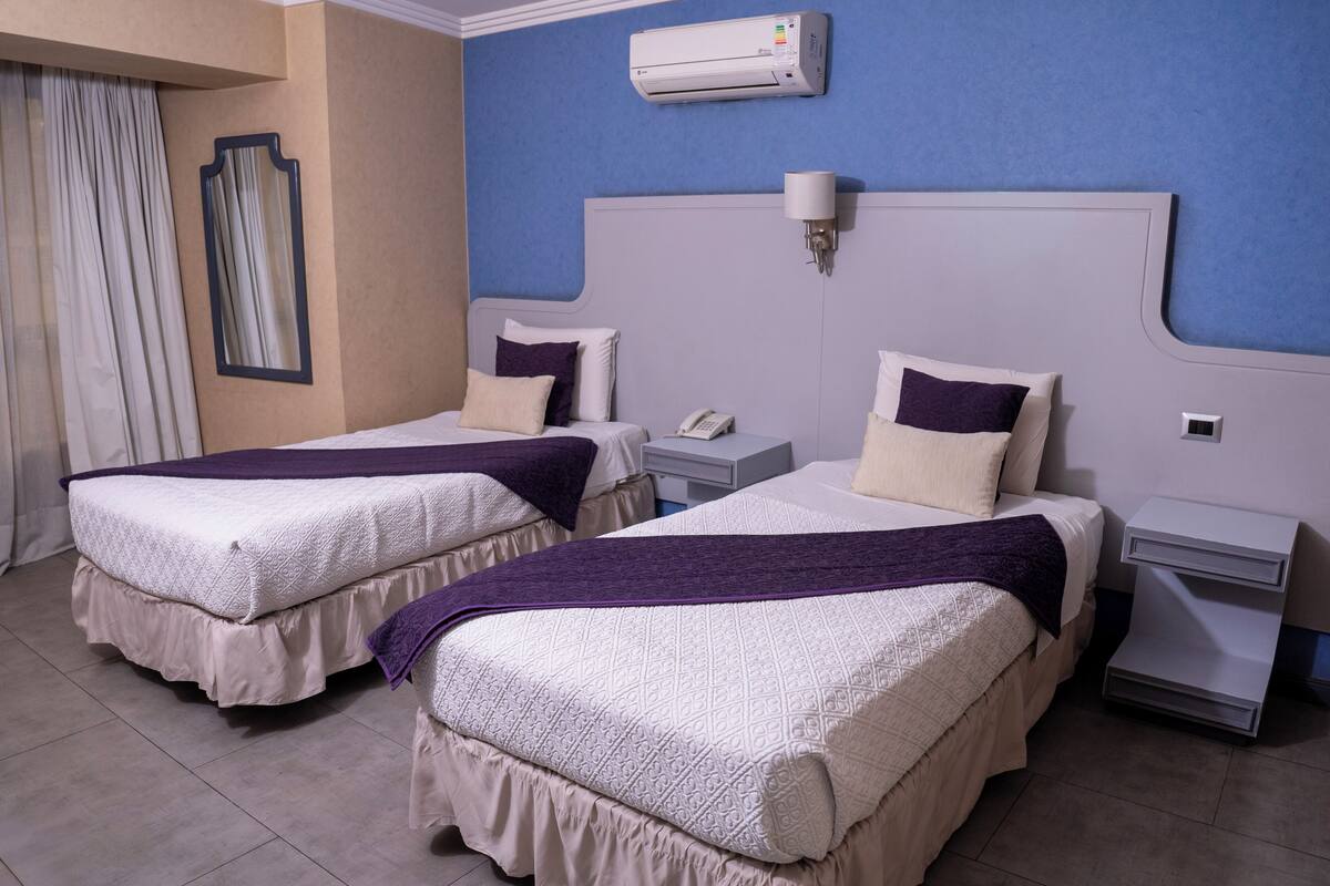 A hotel room with two beds, a nightstand with a phone, a wall-mounted air conditioner, and a window with curtains.