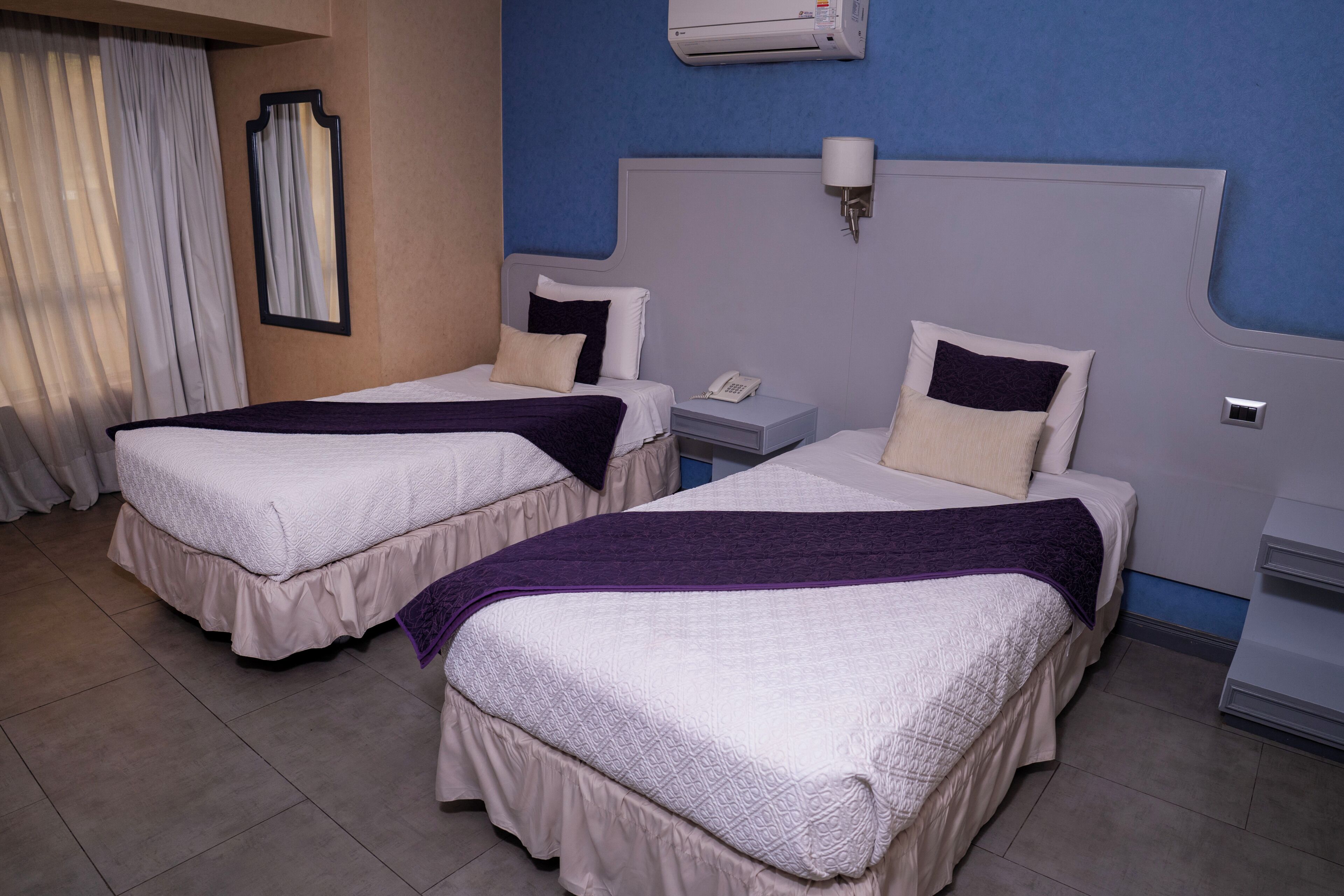 A hotel room with two beds, a nightstand with a phone, a wall-mounted air conditioner, and a window with curtains.