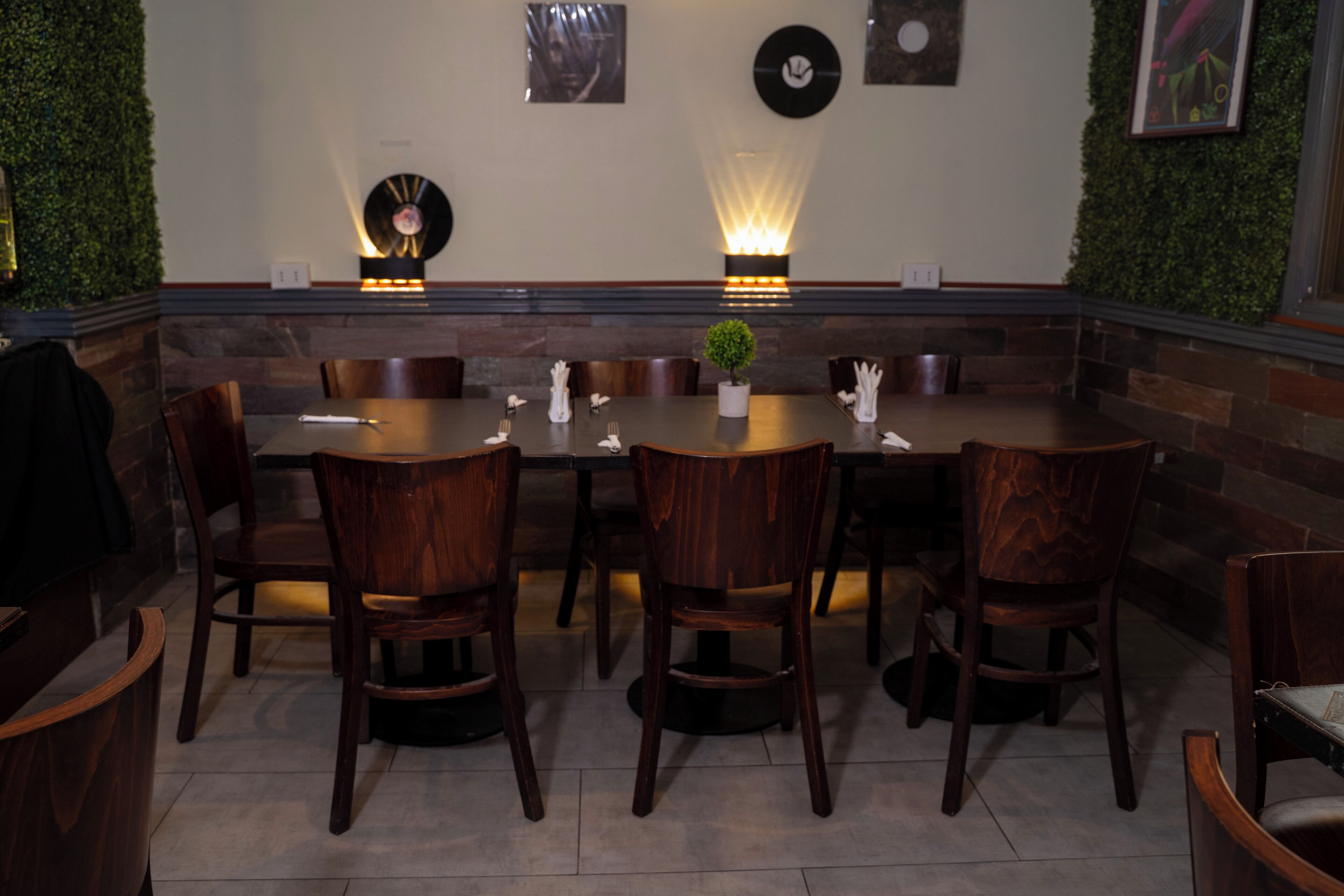 Un espace repas avec une longue table, des chaises en bois, des luminaires fixés au mur et une petite plante en pot.