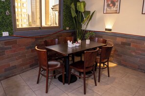 Uma área de refeições com mesa e cadeiras de madeira, um vaso com planta e um candeeiro fixado na parede.